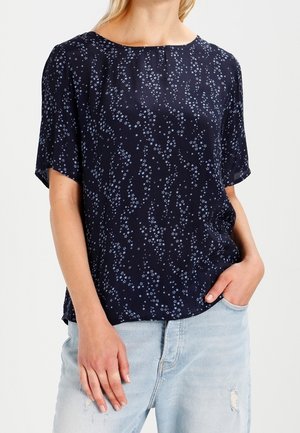 Kobieta ubrana w granatową bluzkę z krótkim rękawem w drobne gwiazdki oraz jasnoniebieskie podarte dżinsy, jedna ręka w kieszeni.