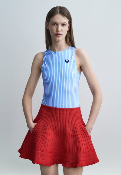 Femme portant un haut sans manches bleu et blanc à rayures avec un logo en forme de cœur et une jupe plissée rouge, mains dans les poches, fond neutre.