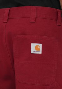 Červené nohavice Carhartt s textúrovanou látkou, so zadnou kapsou ozdobenou bielou značkovou etikou s oranžovým emblémom.