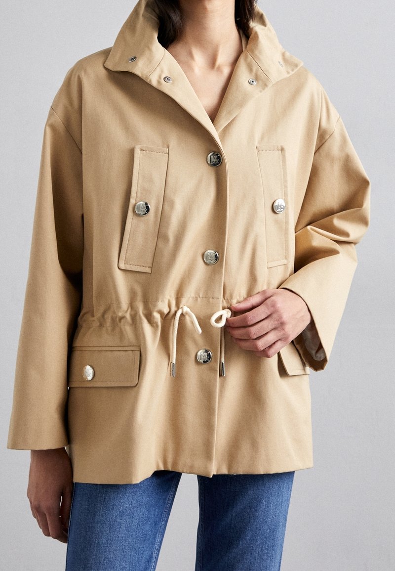 Chaqueta beige con botones plateados en el frente, dos bolsillos en el pecho y dos en la parte inferior, cintura con cordón, combinada con jeans azules sobre un fondo neutro.