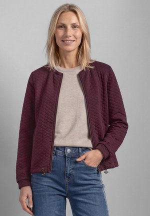 Femme blonde portant une veste matelassée bordeaux par-dessus un pull beige et un jean bleu, une main dans la poche, souriante sur un fond uni.