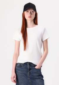 T-shirt bianco a coste con maniche corte, abbinato a una gonna di jeans scura. Indossa un cappellino nero, e il modello ha lunghi capelli rossi.