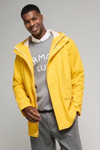 Veste imperméable jaune avec une fermeture éclair à l'avant et des boutons pression, dotée d'une capuche et de poches. Portée sur un sweat-shirt gris avec du texte.