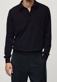 Mężczyzna w czarnym, długim polosie wsuniętym w czarne spodnie z jedną ręką w kieszeni, na neutralnym tle.