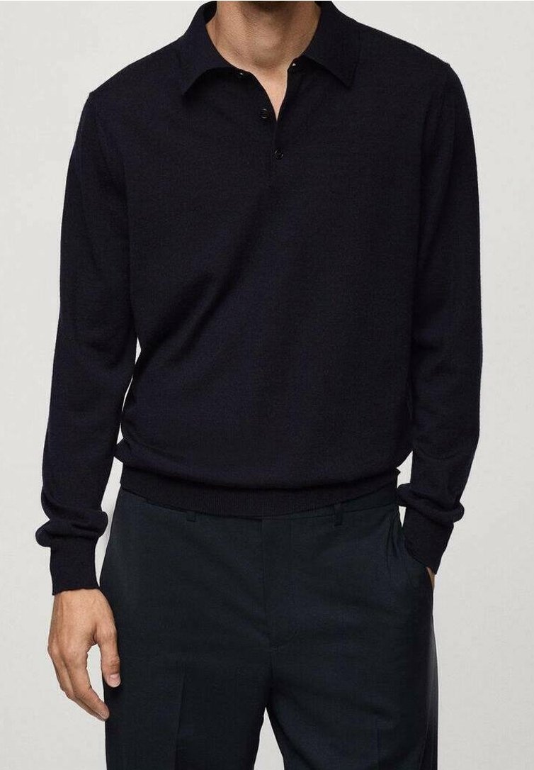 Mężczyzna w czarnym, długim polosie wsuniętym w czarne spodnie z jedną ręką w kieszeni, na neutralnym tle.