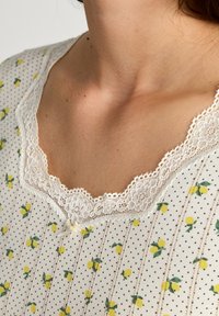 Tessuto bianco con stampa di limoni gialli e piccoli punti neri. Dettaglio del collo in pizzo scallop. Tessitura morbida, design a maniche corte.
