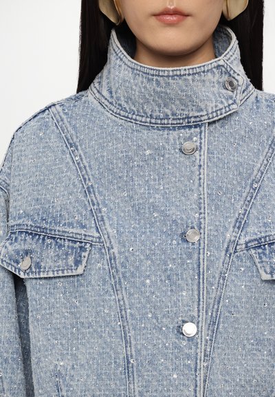 Svijetloplava traper jakna s visokim ovratnikom, srebrnim dugmadi za zakopčavanje i sjajnim detaljima. Ima dva prednja džepa i teksturiranu tkaninu.