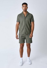 Camicia a maniche corte oversize color verde oliva con bottoni e pantaloni corti abbinati con strisce verticali bianche. Indossato con sneakers bianche e calzini alla caviglia.
