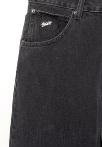 Jean en denim noir montrant une poche avant avec un petit logo blanc brodé et des rivets métalliques aux coins de la poche.