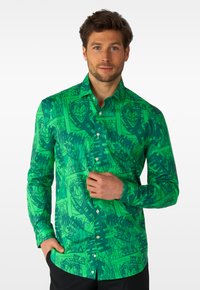 Mann trägt grünes Hemd mit Knöpfen, verziert mit einer dunkelgrünen Joker-Karte und Comic-Mustern, posiert mit einer Hand auf den Knöpfen und der anderen in der Tasche.