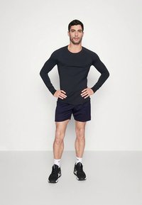 Musta pitkähihaine paita, jossa on pyöreä kaula-aukko, yhdistettynä tummansinisiin shortseihin. Malli seisoo kädet lanteilla, jalassa mustat urheilukengät ja valkoiset sukat.