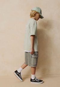 Enfant portant une casquette vert clair et un t-shirt surdimensionné, un short cargo gris, des chaussettes blanches et des baskets noires, marchant devant un fond beige.