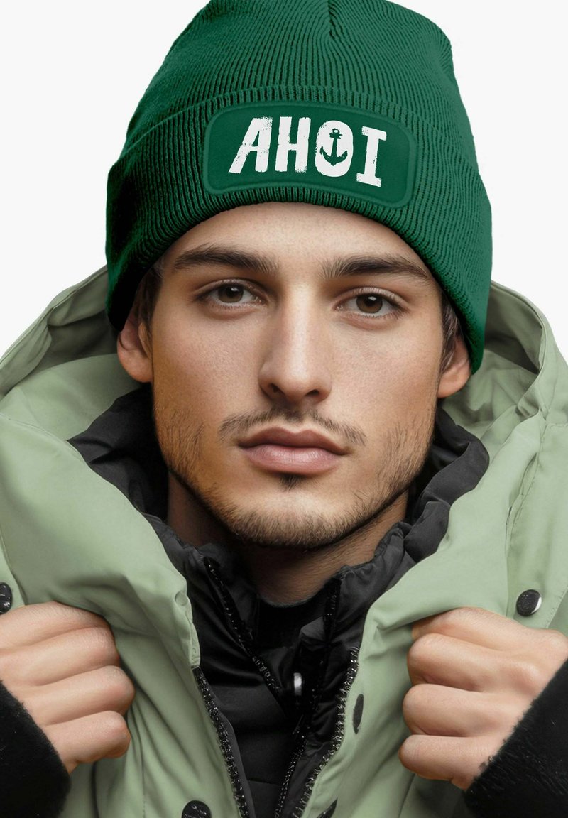 Green knit beanie with "AHOI" and anchor design, paired with a light green zip-up jacket and black inner layer. Soft texture and casual fit.
