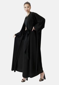 Schwarzes langes Kleid mit weiten Ärmeln, tailliertem Bund und einer Bindeschleife. Weicher, fließender Stoff mit einer glatten, geschmeidigen Textur. Sandalen mit hohen Absätzen.