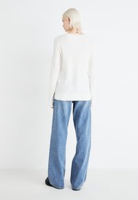 Osoba s platinasto plavom kosom stoji okrenuta leđima, odjevena u bijelu majicu s dugim rukavima, široke plave traperice i crne cipele na jednobojnoj pozadini.