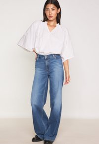 Blusa bianca, ampia e con scollatura a V, maniche corte e larghe; abbinata a jeans blu a vita alta e svasati e scarpe con piattaforma nere.