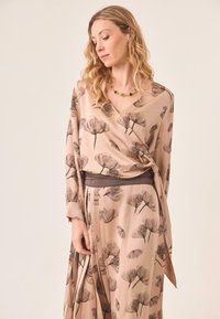 Blusa envolvente fluida en tela beige, con estampados florales oscuros, mangas largas y una cintura ajustada con una banda oscura en contraste.
