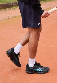 Tennisspieler, der in blauen Shorts, weißen Socken und schwarzen Sportschuhen steht und einen umwickelten Schlägergriff in der Hand hält, während er auf einem roten Sandplatz steht.