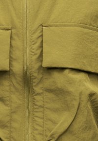 Κοντινή λήψη του μπροστινού μέρους ενός χακί σακακιού που δείχνει το φερμουάρ και δύο τσέπες με καπάκι με ορατές λεπτομέρειες ραφής.