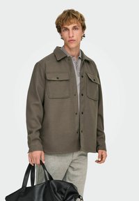 Chemise extérieure marron tissée avec deux poches poitrine, boutonnée sur le devant, avec un col, associée à une chemise grise et un pantalon léger à carreaux. Sac noir à la main.