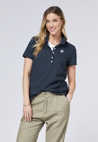 Navyblaues Polohemd mit Kragen, ausgestattet mit einem kleinen weißen Pferdelogo, fünf Knöpfen und kurzen Ärmeln. Kombination mit hellbeigen Hosen.