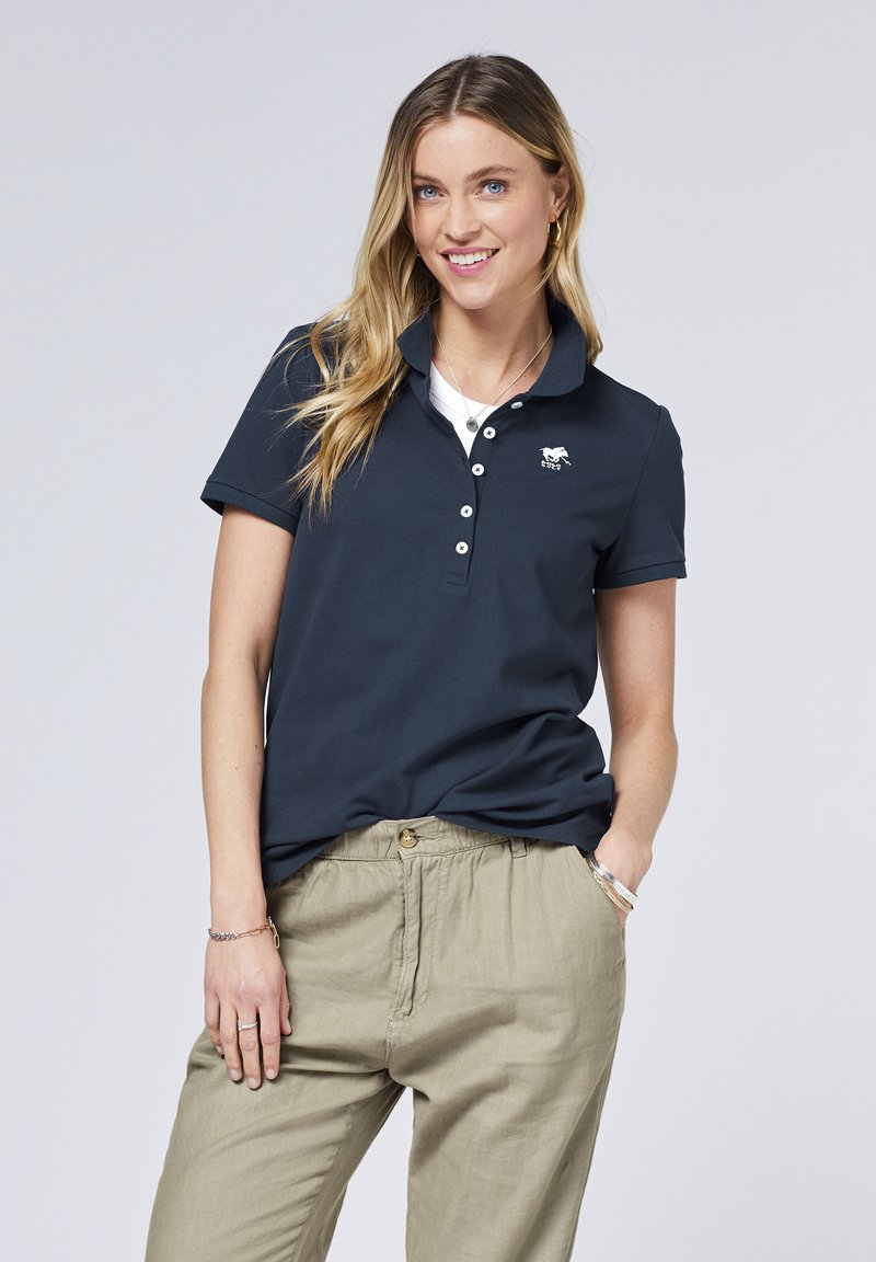 Navyblaues Polohemd mit Kragen, ausgestattet mit einem kleinen weißen Pferdelogo, fünf Knöpfen und kurzen Ärmeln. Kombination mit hellbeigen Hosen.