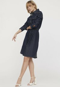 Robe chemise en denim bleu foncé, dotée d'un col, d'une fermeture boutonnée sur le devant et d'une taille cintrée. Ceinturée avec des fentes sur les côtés. Finitions avec des accents dorés.