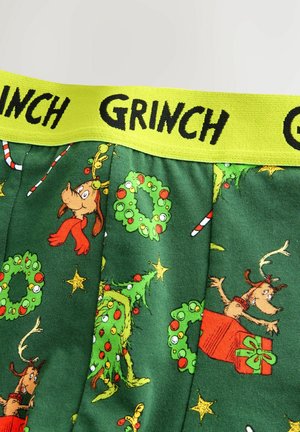 Grüner Stoff mit Grinch-weihnachtlichen Mustern, darunter Kränze, Sterne, Zuckerstangen und ein Hund mit Geweih, gelber Bund mit der Aufschrift "GRINCH".