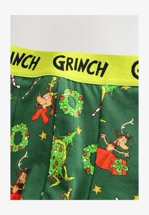 Grüner Stoff mit Grinch-weihnachtlichen Mustern, darunter Kränze, Sterne, Zuckerstangen und ein Hund mit Geweih, gelber Bund mit der Aufschrift "GRINCH".
