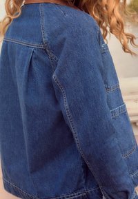 Dżinsowa kurtka w ciemnoniebieskim kolorze, z luźnym krojem, długimi rękawami, plisą z tyłu i kontrastowym przeszyciem na kieszeniach.