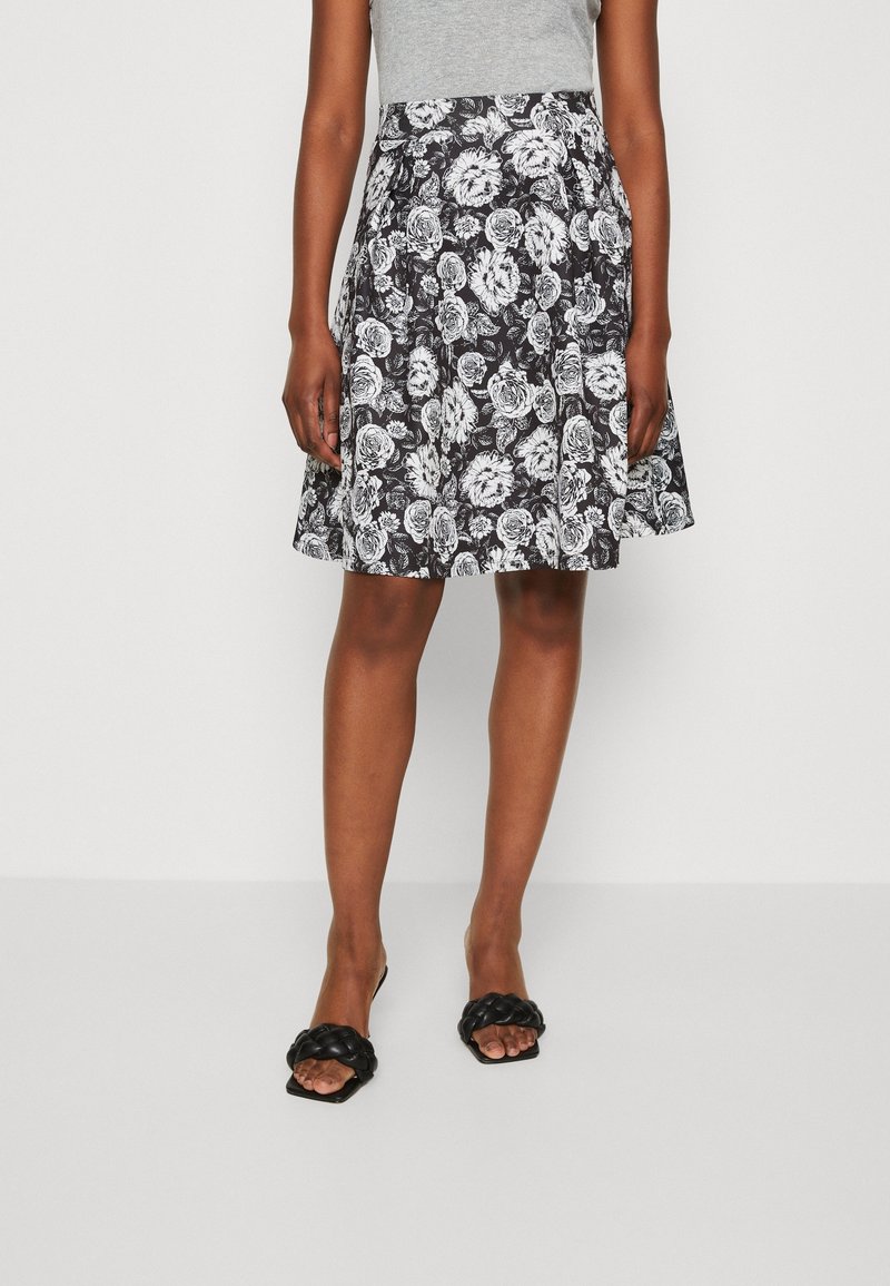 Femme portant une jupe noire et blanche à fleurs mi-longue, un top sans manches gris clair et des sandales noires tressées, debout devant un fond blanc.