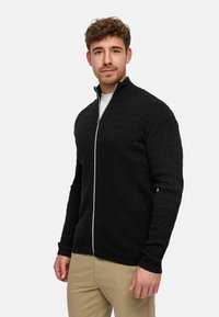 Svart stickad tröja med dragkedja och texturerad kabelstickning, ribbad krage och muddar, kombinerad med en vit understickad tröja och beige byxor.