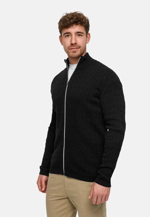 Sort strikket trøje med lynlås og et tekstureret kabelflet-design, med ribbet krave og manchetter, kombineret med en hvid undertrøje og beige bukser.