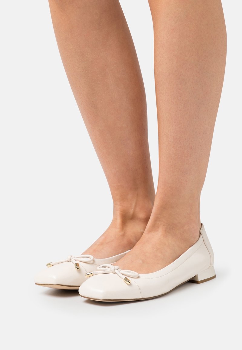Femme portant des ballerines en cuir blanc à talons bas et petits nœuds décoratifs avec des embouts dorés sur un fond clair.
