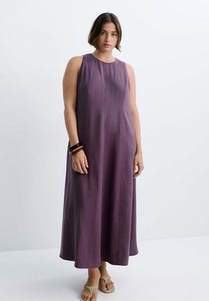 Mujer con cabello corto y oscuro, vestida con un vestido largo morado sin mangas y sandalias beige, de pie frente a un fondo blanco liso.