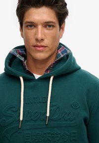 Teal hoodie met aantrekkoorden, voorzien van een gestructureerd logo en een geruite flanelvoering die zichtbaar is bij de kraag. Zacht stof met een casual ontwerp.