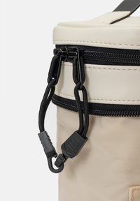 Sac cylindrique beige avec une fermeture éclair noire, finition texturée et une corde détachable avec un clip en plastique. Design minimaliste avec des couleurs contrastées.