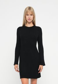 Donna con capelli biondi lisci che indossa un abito a maniche lunghe nero aderente a costine, in piedi davanti a uno sfondo bianco semplice.