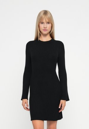Donna con capelli biondi lisci che indossa un abito a maniche lunghe nero aderente a costine, in piedi davanti a uno sfondo bianco semplice.
