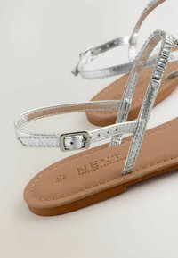 Zilveren sandalen met banden, voorzien van metalen straps, met een zachte bruine voetbed en een verstelbare gesp. Het ontwerp bevat meerdere dunne banden.