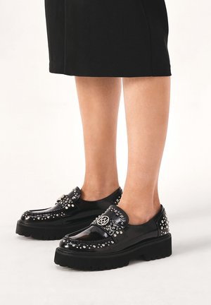Ben iført sort nederdel og sorte platform loafers med nitter samt sølv logo på fronten og hælen.