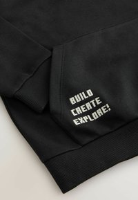 Sudadera negra con puños acanalados; presenta el texto blanco "¡CONSTRUYE CREA EXPLORA!" bordado en la manga; textura suave y lisa.