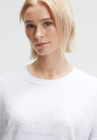 Junge Frau mit kurzem blonden Haar und blauen Augen, die ein weißes Sweatshirt mit schwachem Text "Rise Up" und motivierenden Sprüchen trägt.