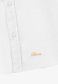 Hvid skjorte med knapper lavet af et tekstureret stof. Har et gult broderet logo "Blkovi" på nederste venstre side. Klassisk kravedesign.