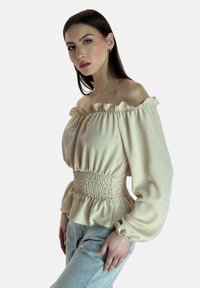 Off-Shoulder-Bluse in hellem Beige mit Rüschenausschnitt, smockiertem Bund und voluminösen Ärmeln, kombiniert mit hellblauen Jeans.