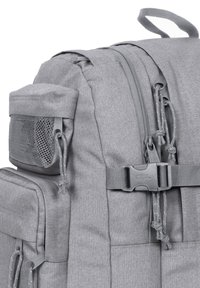 Mochila de tela gris que cuenta con múltiples compartimentos, un bolsillo lateral de malla, cierres de cremallera y un detalle de correa con hebilla.