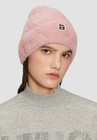 Różowa, puszysta czapka z zaokrąglonym wierzchem, z małym prostokątnym logo. Miękka tekstura kontrastuje z szarym swetrem z wysokim kołnierzem.