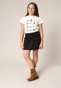 Witte T-shirt met bedrukte graphics, zwarte suède rok met knopen aan de voorkant en zakken, bruine enkellaarsjes, staand op een neutrale achtergrond.