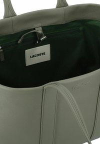 Borsa tote in pelle grigia chiara con interno verde, tasca con zip e etichetta Lacoste. Presenta manici robusti e un'esterno testurizzato.
