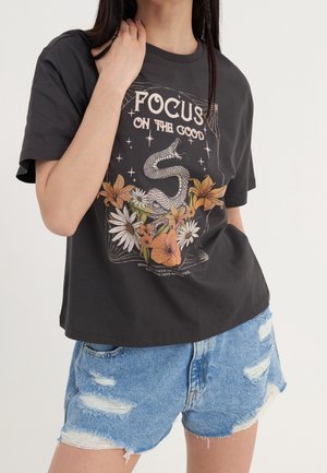 Osoba nosząca czarną koszulkę z napisem "Focus on the Good", wąż i kwiatowy wzór, w połączeniu z podartymi niebieskimi szortami jeansowymi.